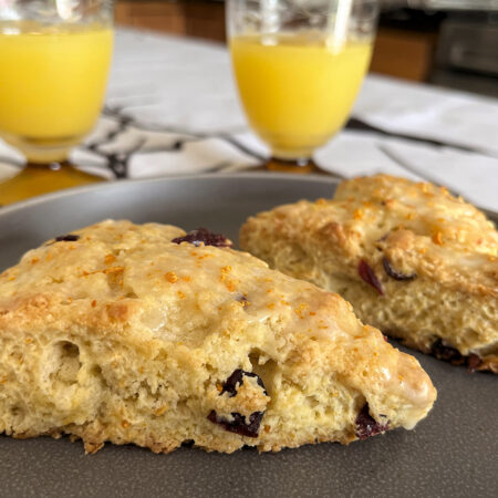 Two cranberry orange scones with an orange glaze on a gray plate over a white tablecloth with a bare branch design with two glasses of orange juice