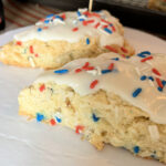 Two coconut scones with red white and blue sprinkles topped with an opaque vanilla white glaze and red, white ,and blue sprinkles on a white cake board. with a portion of a vanilla extract bottle next to it.
