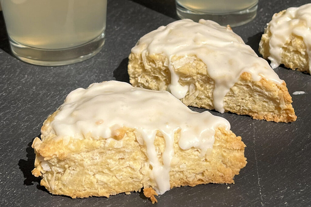 Two coconut scones covered and dripping with an almond flavored white glaze on a black slate server and two glasses of lemonade behind them.