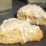 Two coconut scones covered and dripping with an almond flavored white glaze on a black slate server and two glasses of lemonade behind them.