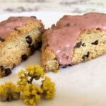 Two triangular scones with chocolate chips with a red raspberry glaze and yellow tree flowers next to them on a white plate sitting on a white tablecloth with spring lilac flowers and green leaves.