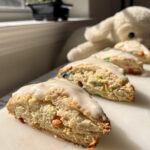 Three scones filled with mult-colored mini M&Ms on a concrete bench with a stuffed lamb in the background.