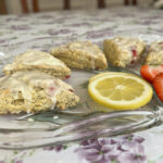 Five triangular strawberry lemon poppy seed scones with bits of strawberries and dried lemon zest on top of a light white on a glass flowered tray on a white tablecloth with lavender flowers and green leaves.